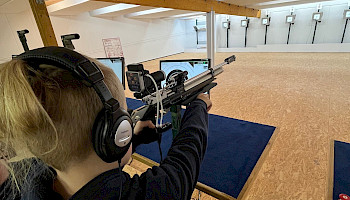 Ein Junge sitzt mit Kopfhörern an einem Schiessstand. Man sieht wie er mit einem Gewehr auf eine entfernte Zielscheibe zielt.